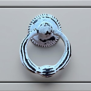 Könnte beinhalten: Ein runder Ringgriff aus Metall mit einem abgenutzten weißen Finish auf schwarzer Basis. Das Beschlagteil verfügt über eine runde Montageplatte mit geriffeltem Rand und einen beweglichen Ring mit texturierter, abgenutzter Optik auf einer Fläche.