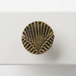 Puede incluir: Un pomo de mueble circular con acabado en latón antiguo que presenta un patrón de concha en forma de abanico con relieve y textura. La superficie metálica tiene una pátina oscura y desgastada en las ranuras, resaltando el diseño.