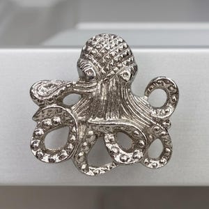 May include: A silver-toned metal cabinet knob shaped like an octopus. The octopus features a textured, rounded head with a cross-hatched pattern and eight curved tentacles with visible circular suction cup details along the underside of each arm.