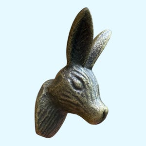 May include: A bronze-colored wall-mounted rabbit head sculpture. The sculpture features detailed fur texture and long, upright ears. The rabbit's head is angled to the right, with a detailed nose and eye.