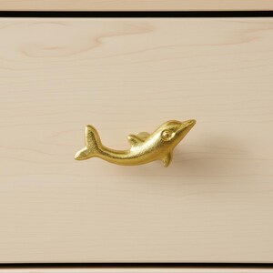 Puede incluir: Un tirador de armario dorado con forma de delfín. El tirador está montado en un cajón de madera de color claro. El delfín mira hacia la derecha, con la cola y las aletas claramente visibles. El tirador añade un toque náutico a los muebles.