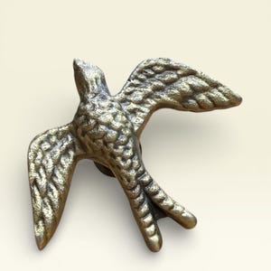 Può includere: Un battente per porta a forma di uccello, color bronzo. Il battente raffigura un uccello in volo, con dettagli di piume sulle ali e sul corpo. L'uccello è montato su una base circolare, pronto per essere fissato a una porta.