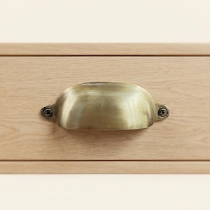 Peut inclure: Une poignée de tiroir en forme de coquille en laiton avec une finition brossée, fixée sur une façade de tiroir en bois clair avec deux vis métalliques visibles sur les côtés gauche et droit, offrant une prise fonctionnelle pour ouvrir le tiroir.