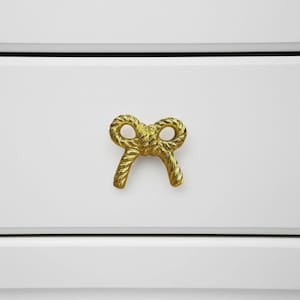 Pode incluir: Um puxador de armário dourado em forma de laço. O puxador de metal tem um design texturizado semelhante a uma corda. É montado em uma porta de armário branca. O laço está centrado na porta.