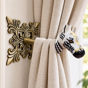 May include: A curtain tieback featuring a wall-mounted antique brass-toned metal base with an ornate, square, leaf-like design. A horizontal rod extends from the center, ending in a ceramic zebra head finial with black stripes and gold accents.