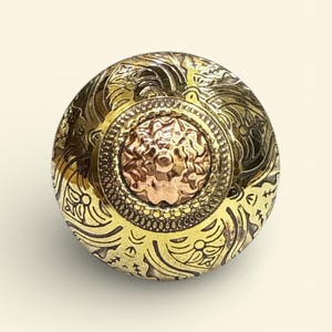 Puede incluir: Perilla de armario redonda y ornamentada, con acabado en latón y cobre. La perilla presenta detallados patrones florales y de volutas. El centro tiene un medallón de color cobre con un diseño floral. Elemento de ferretería decorativo.