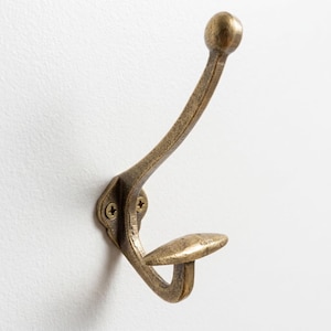 May include: A wall-mounted coat hook with a distressed antique brass finish. The hook features a curved upper arm ending in a rounded knob and a shorter, horizontal lower prong. It is secured to the wall with two visible brass-colored screws.