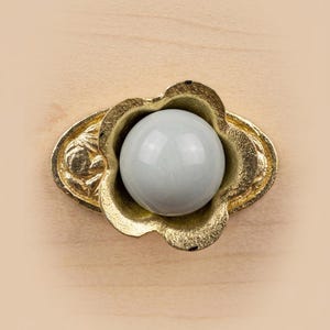 Peut inclure: Un bouton de tiroir doté d'une pierre sphérique centrale, lisse et gris pâle, insérée dans une base en métal doré texturé en forme de fleur à cinq pétales. La base métallique possède une plaque arrière ovale avec un motif de feuilles en relief.