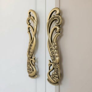 May include: Two ornate, antique-style brass door handles. The handles feature a peacock design with intricate detailing and a warm, golden hue. They are mounted on a light-colored, neutral background.