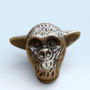 Könnte beinhalten: Ein bronzefarbenes, affengesichtiges Dekorationsobjekt mit detaillierten Gesichtszügen, einschließlich Augen, Nasenlöchern und Ohren. Die Skulptur hat eine strukturierte Oberfläche.
