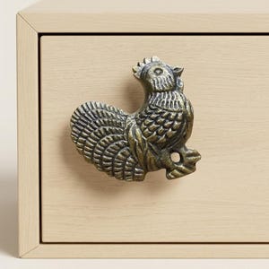 May include: A brass-toned metal cabinet knob shaped like a rooster in profile, featuring detailed feather textures and a dark antique finish, mounted on a light-colored wood drawer front with a smooth, horizontal grain pattern.