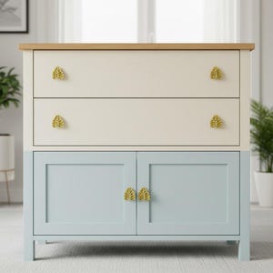 May include: A two-tone cabinet. The lower section is light blue, the upper section is off-white. It has two drawers and two doors, each with gold leaf-shaped handles. The top is light wood. A stylish storage solution.