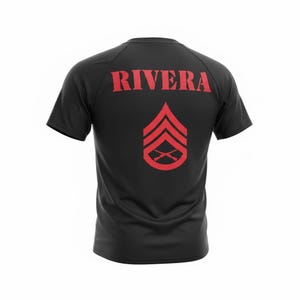 May include: Black short-sleeved t-shirt with the name "RIVERA" printed in red across the shoulders. Below the name is a red insignia featuring three chevrons and crossed rifles, commonly associated with military ranks.