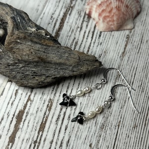 May include: A pair of silver earrings with black shark tooth charms and small white pearls. The earrings are displayed on a white wooden surface with a piece of driftwood and a seashell in the background.