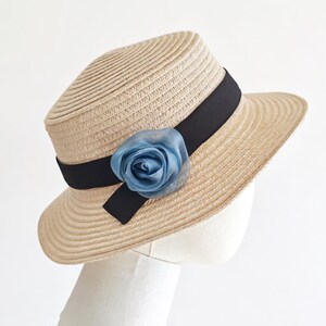 May include: A beige straw hat with a black band and a light blue fabric rose. The hat has a rounded crown and a wide brim. The hat is displayed on a white mannequin head.