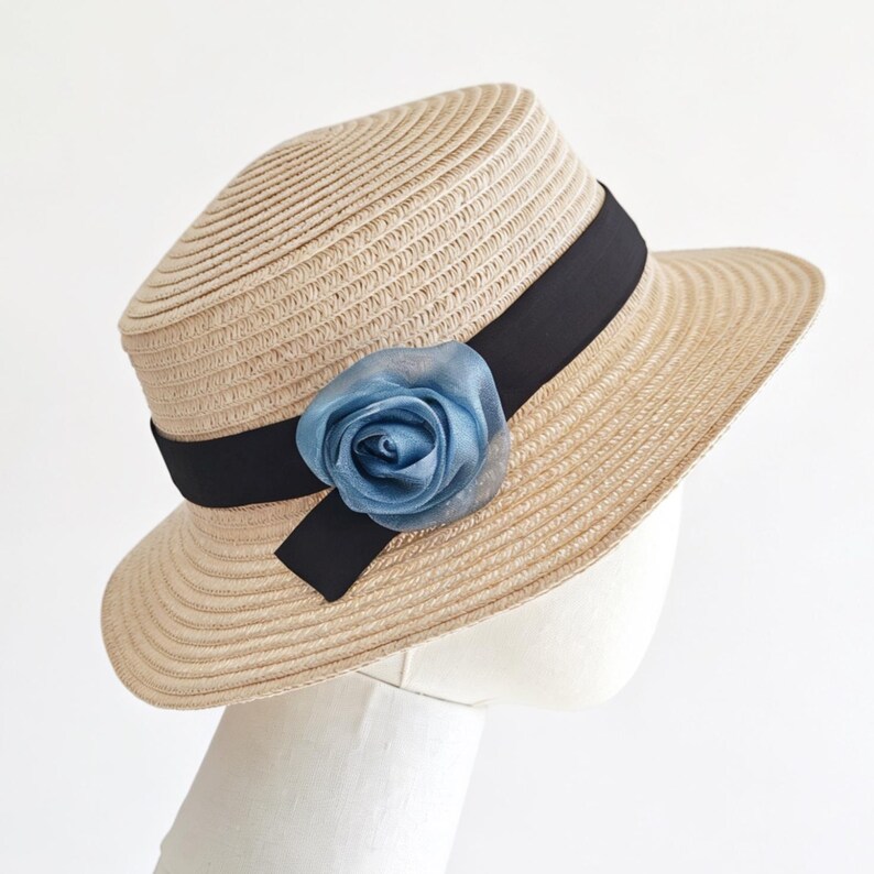 May include: A beige straw hat with a black band and a light blue fabric rose. The hat has a rounded crown and a wide brim. The hat is displayed on a white mannequin head.