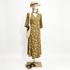 May include: A mannequin displays a floral print dress, straw hat, earrings, and a woven tote bag. The olive green dress features a V-neck and short sleeves. Sandals with a floral pattern are at the base of the display.