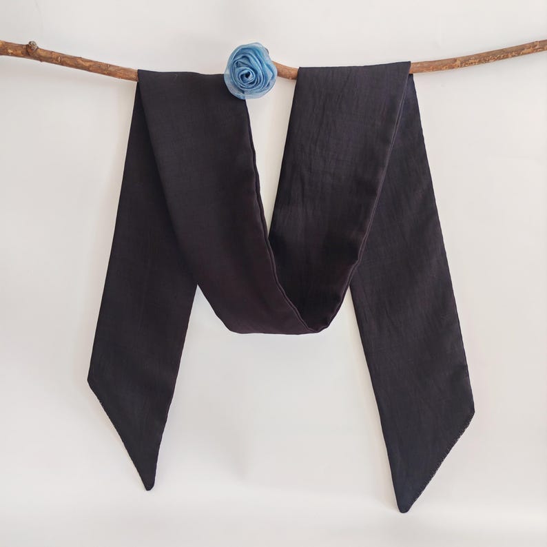 May include: A long, dark charcoal-colored scarf with pointed ends, draped over a thin, brown branch. A small, light blue fabric flower is attached to the branch. The scarf is made of a soft, flowing material.