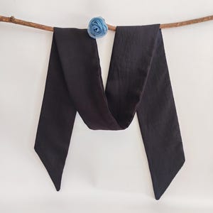 May include: A long, dark charcoal-colored scarf with pointed ends, draped over a thin, brown branch. A small, light blue fabric flower is attached to the branch. The scarf is made of a soft, flowing material.