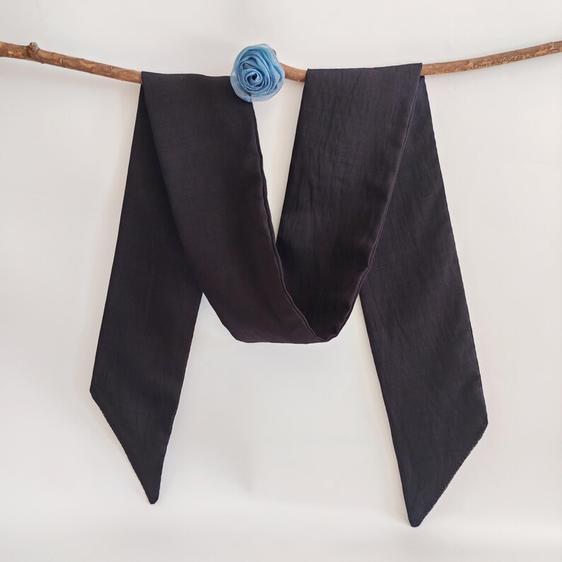 May include: A long, dark charcoal-colored scarf with pointed ends, draped over a thin, brown branch. A small, light blue fabric flower is attached to the branch. The scarf is made of a soft, flowing material.