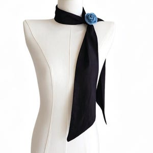 May include: A black neck scarf with a light blue rose accent. The scarf is draped on a mannequin, showcasing its length and design. The rose is positioned near the neck, adding a touch of color and detail to the accessory.