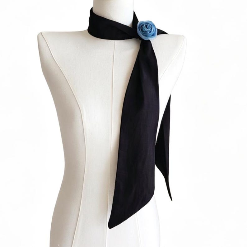 May include: A black neck scarf with a light blue rose accent. The scarf is draped on a mannequin, showcasing its length and design. The rose is positioned near the neck, adding a touch of color and detail to the accessory.
