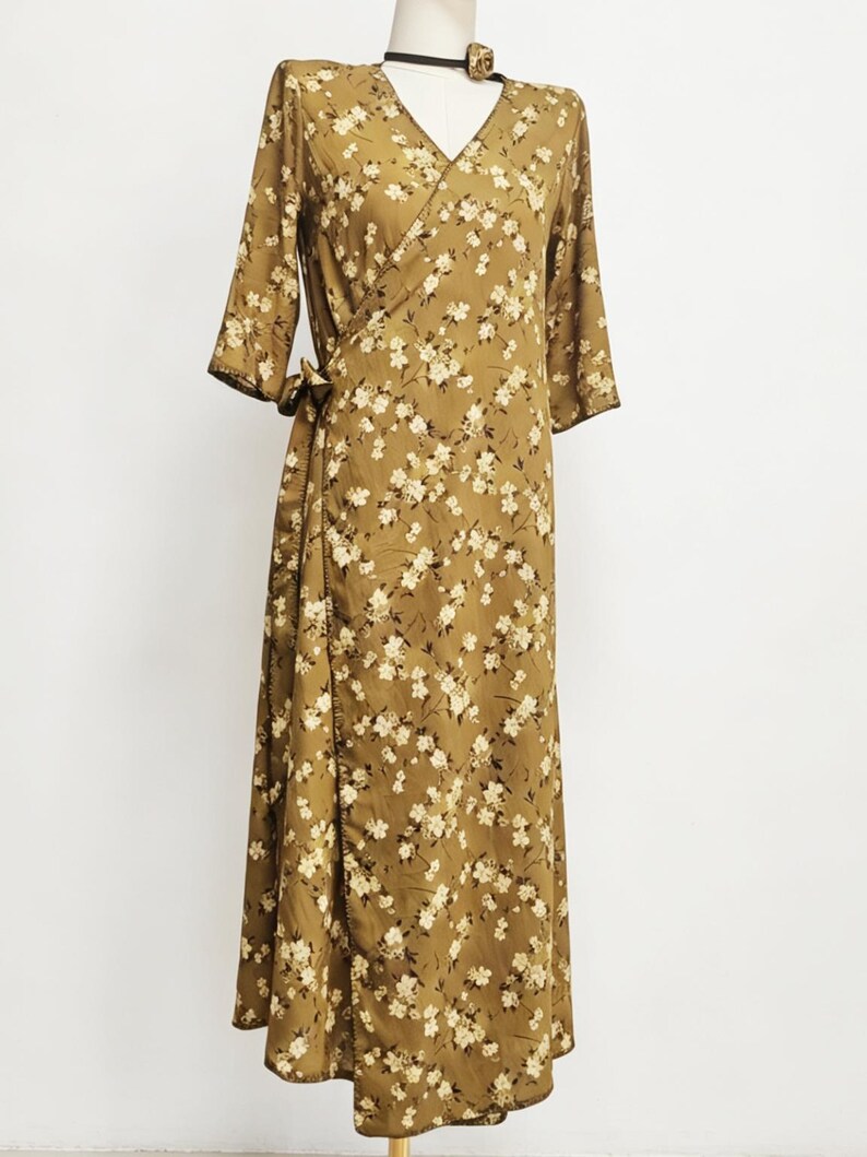 May include: A long, olive-green wrap dress with a floral pattern. The dress features a V-neck, three-quarter sleeves, and a tie closure at the waist. The fabric is covered in small, white and cream-colored flowers.