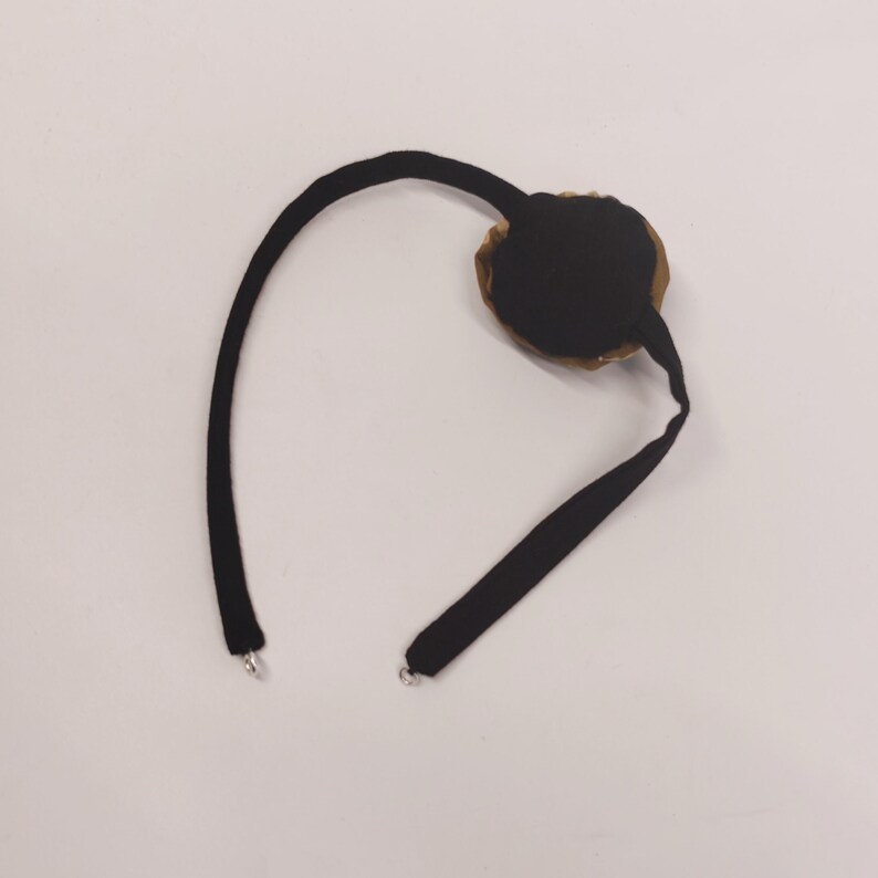 May include: A black eye patch with a circular, brown and black design. The patch is connected to a black strap that curves around and has two silver metal clasps.