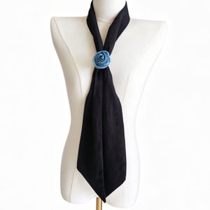May include: A black scarf with a light blue rose accent, displayed on a white mannequin. The scarf is draped and knotted, with the rose positioned at the knot. The scarf's fabric appears soft and flowing, creating a simple yet elegant look.