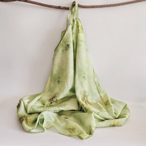 May include: A light green tote bag with a floral pattern, hanging from a wooden branch. The bag is made of a soft, flowing fabric and features a stitched edge. The bag's straps are also made of the same fabric.