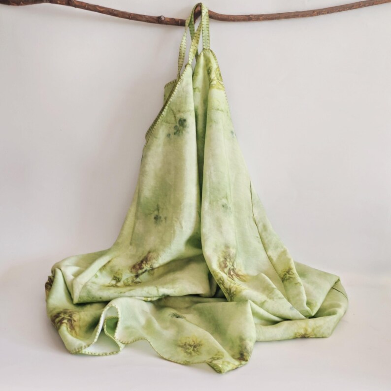 May include: A light green tote bag with a floral pattern, hanging from a wooden branch. The bag is made of a soft, flowing fabric and features a stitched edge. The bag's straps are also made of the same fabric.