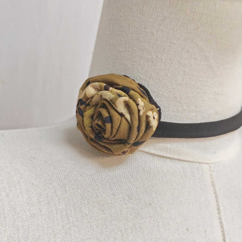 May include: A fabric rose in shades of olive green, tan, and navy blue, attached to a black elastic choker. The floral accent is made of patterned fabric, creating a unique accessory.