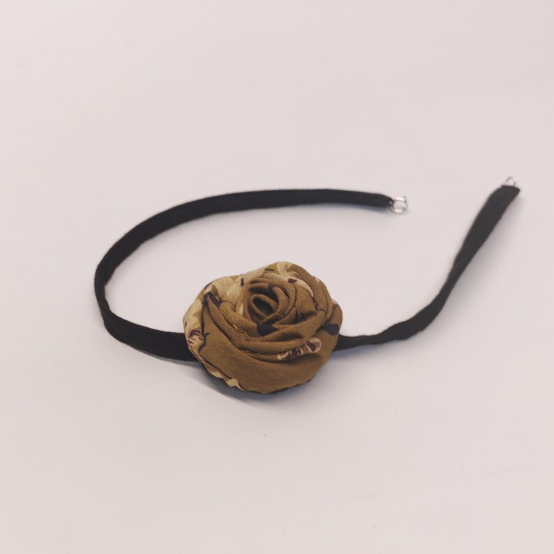 May include: A black choker necklace with a large, fabric rose in shades of brown and tan. The rose is the focal point, and the necklace has a simple, elegant design. The choker is made of a soft, black material.