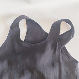 May include: Close-up of a black tank top with wide straps. The top is made of a lightweight fabric and has a simple, minimalist design. The straps cross in the back, creating a unique silhouette. The top is likely for casual wear.