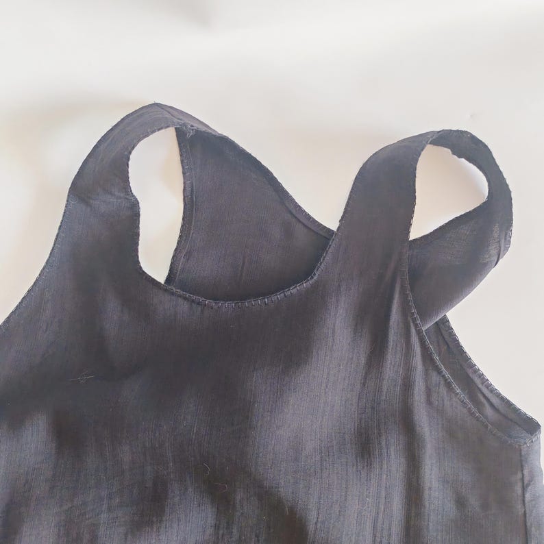 May include: Close-up of a black tank top with wide straps. The top is made of a lightweight fabric and has a simple, minimalist design. The straps cross in the back, creating a unique silhouette. The top is likely for casual wear.