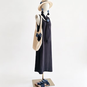 May include: A mannequin displays a navy blue dress, straw hat, earrings, and sandals, all adorned with blue bows. A cream-colored, open-weave tote bag with a blue bow hangs from the mannequin's shoulder. The ensemble suggests a summer fashion theme.