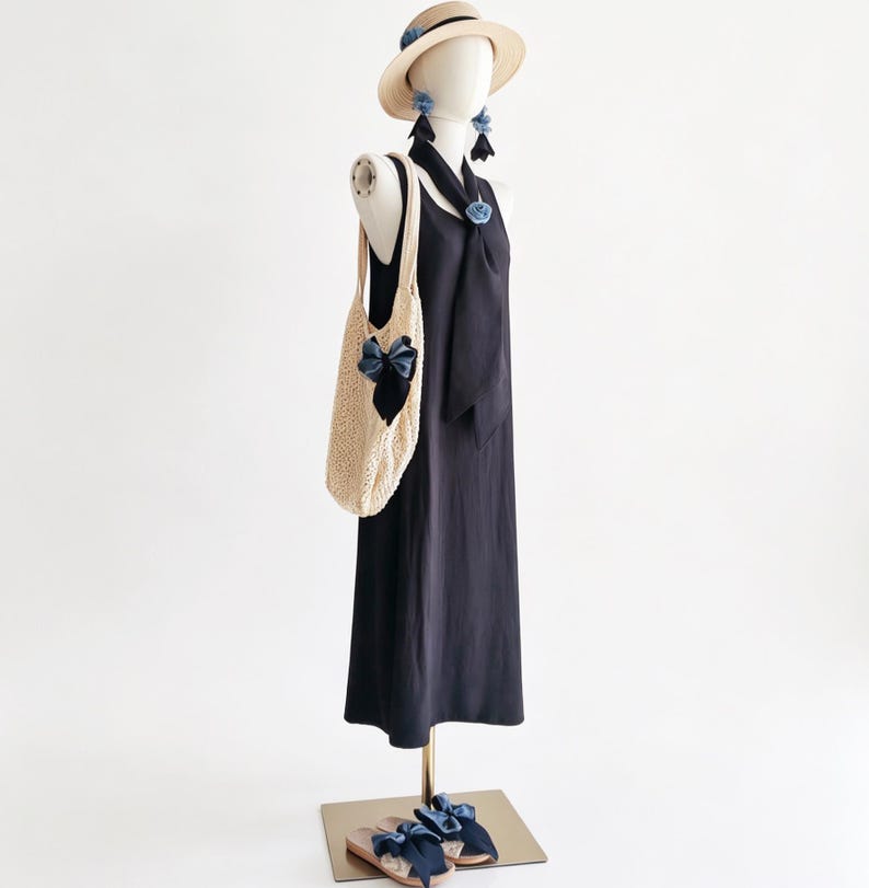 May include: A mannequin displays a navy blue dress, straw hat, earrings, and sandals, all adorned with blue bows. A cream-colored, open-weave tote bag with a blue bow hangs from the mannequin's shoulder. The ensemble suggests a summer fashion theme.