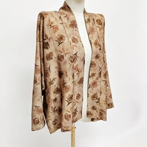 K&ouml;nnte beinhalten: Eine hellbraune Jacke mit offener Vorderseite und einem sich wiederholenden dunkelbraunen Blumenmuster. Das Kleidungsst&uuml;ck hat lange &Auml;rmel, einen breiten Kragen und einen geraden Saum und wird auf einer Schneiderpuppe pr&auml;sentiert.