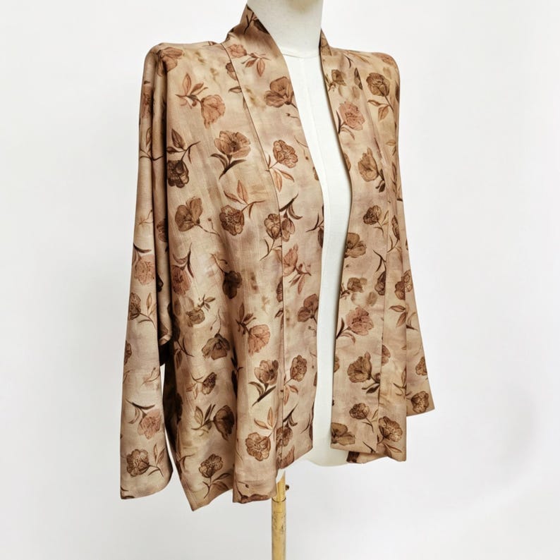 K&ouml;nnte beinhalten: Eine hellbraune Jacke mit offener Vorderseite und einem sich wiederholenden dunkelbraunen Blumenmuster. Das Kleidungsst&uuml;ck hat lange &Auml;rmel, einen breiten Kragen und einen geraden Saum und wird auf einer Schneiderpuppe pr&auml;sentiert.
