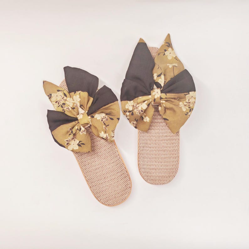 May include: A pair of beige woven slippers with large decorative bows. The bows are a combination of dark brown and floral patterned fabric in shades of yellow and white. The slippers are presented on a white background.