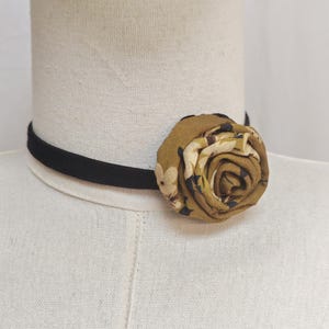 May include: A black choker necklace with a floral rose accent. The rose is made of fabric with a pattern of olive green, cream, and navy blue. The choker is displayed on a white mannequin.