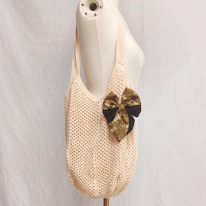 May include: A cream-colored, open-weave tote bag with a large bow. The bow is a combination of black and a floral print in shades of green and brown. The bag has a shoulder strap and is displayed on a mannequin.