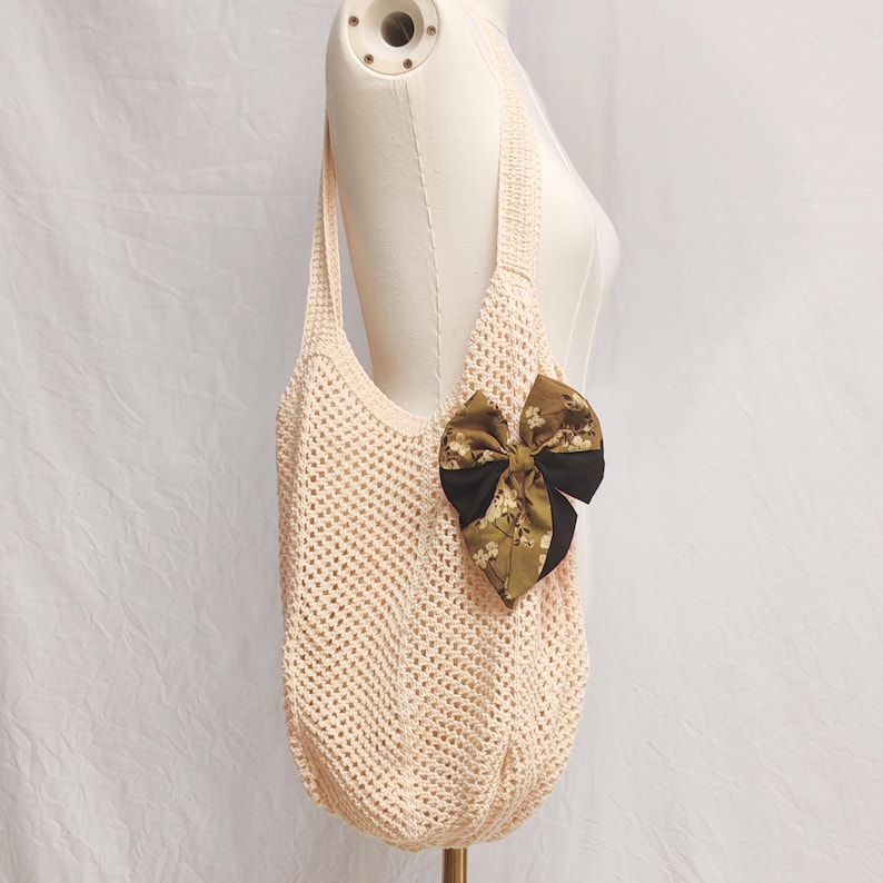 May include: A cream-colored, open-weave tote bag with a large bow. The bow is a combination of black and a floral print in shades of green and brown. The bag has a shoulder strap and is displayed on a mannequin.