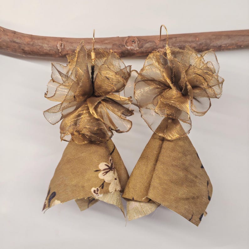 May include: A pair of handmade gold earrings. Each earring features a layered design with a sheer, ruffled top and a bell-shaped bottom. The bottom portion is a patterned fabric with white floral accents.