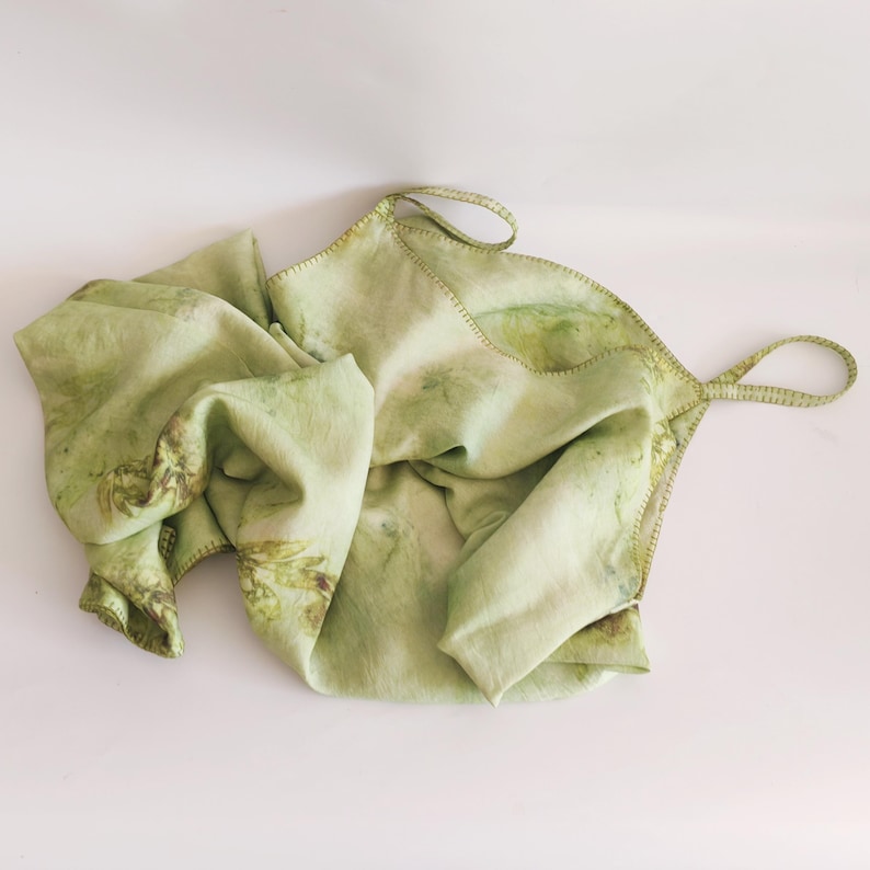 May include: A light green, handmade textile with a botanical print. The fabric is folded and has a stitched edge with two loop handles. The textile has a natural, organic appearance, with variations in color and pattern.