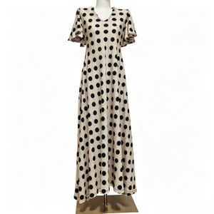 May include: A floor-length beige dress featuring a repeating pattern of large black polka dots, a V-neckline, and short flutter sleeves. The garment has a fitted bodice that transitions into a flowing, A-line skirt that reaches the floor.