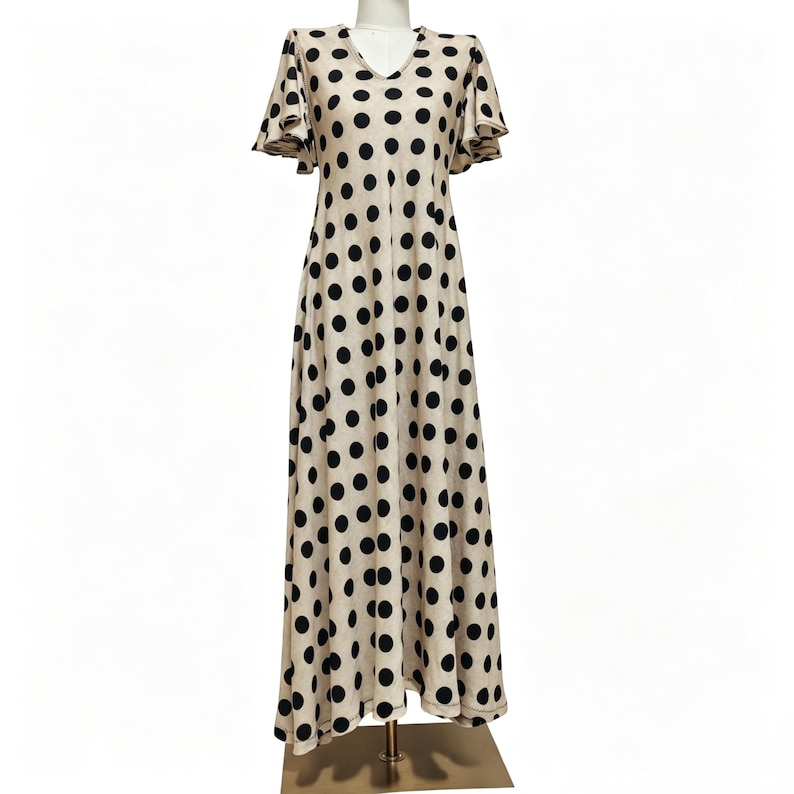 May include: A floor-length beige dress featuring a repeating pattern of large black polka dots, a V-neckline, and short flutter sleeves. The garment has a fitted bodice that transitions into a flowing, A-line skirt that reaches the floor.