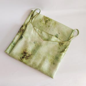 May include: A light green camisole with thin straps, featuring a botanical print in shades of green and brown. The edges are stitched with a contrasting thread. The garment is folded on a white surface.