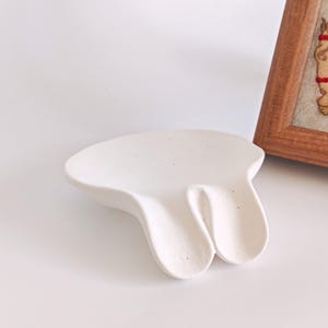 May include: A white, organically shaped ceramic dish with a textured surface. The dish has a unique design with two curved sections. A wooden framed artwork is partially visible in the background.