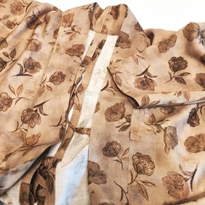 K&ouml;nnte beinhalten: Ein hellbrauner Morgenmantel im Kimono-Stil mit einem sich wiederholenden dunkelbraunen Blumenmuster. Das Kleidungsst&uuml;ck hat weite &Auml;rmel, einen strukturierten Kragen und d&uuml;nne Stoffb&auml;nder an den Vorderteilen zum Verschlie&szlig;en, mit weichem Fall.